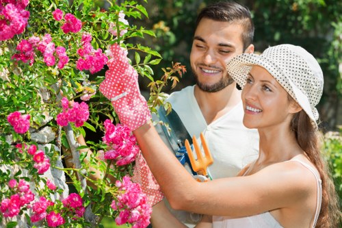A woman and a man are engaged in gardening activities in a lush, well-maintained garden. The woman, wearing a white hat with black polka dots and pink gardening gloves, is pruning or trimming a vibrant pink flowering shrub using her hands. The man, standing close beside her, is smiling and observing her work; he has short dark hair and a beard, and is dressed casually. In the background, there are various green foliage, including trees and shrubs, contributing to a dense, healthy outdoor environment. The scene suggests a sunny day with natural light highlighting the vivid colours of the flowers and greenery. The garden features a mix of flowering plants, a neatly trimmed lawn, and possibly garden tools, indicating routine outdoor maintenance. This outdoor space exemplifies typical features of a Camberwell garden, including diverse plantings and structured garden layout, aligning with professional gardening and landscaping standards that Garden Maintenance Camberwell offers to local homeowners.
